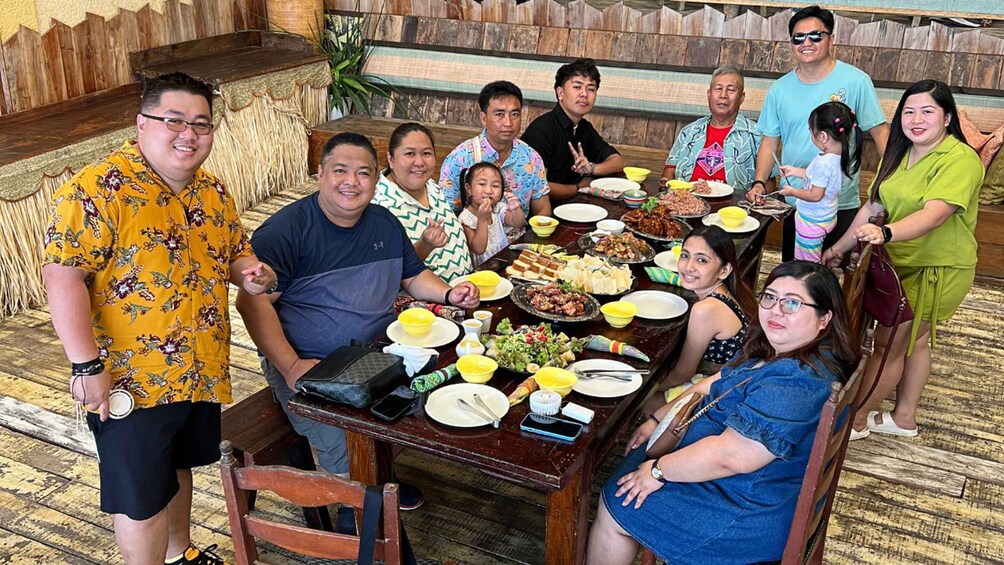 A group of people seated around a table with various dishes, in a room with wooden walls and a thatched ceiling. 2