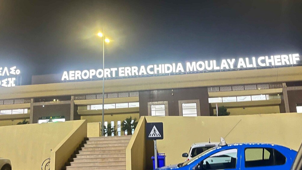 The exterior of the Aeroport Errachidia Moulay Ali Cherif airport with a sign, stairs, and a blue car parked in front. 4