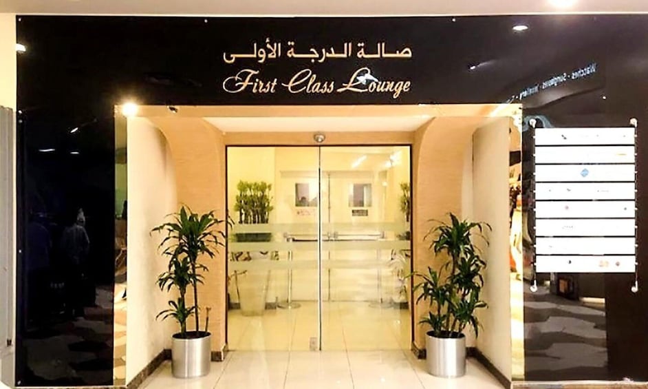 A building entrance with a sign reading 'First Class Lounge' and potted plants. 1