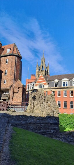 A historic stone structure with a red-tiled roof, a brick tower, and a Gothic-style building with spires in the background. 4