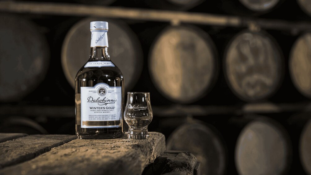 A bottle of Dalwhinnie Winter's Gold whisky and a glass on a wooden surface with barrels in the background. 3