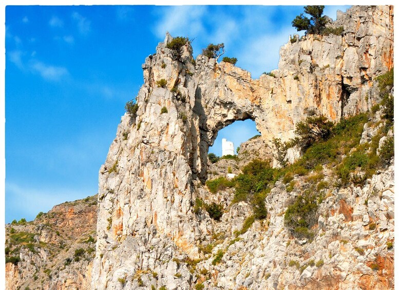 A rocky cliff with a natural arch formation and a white building at the base. 5