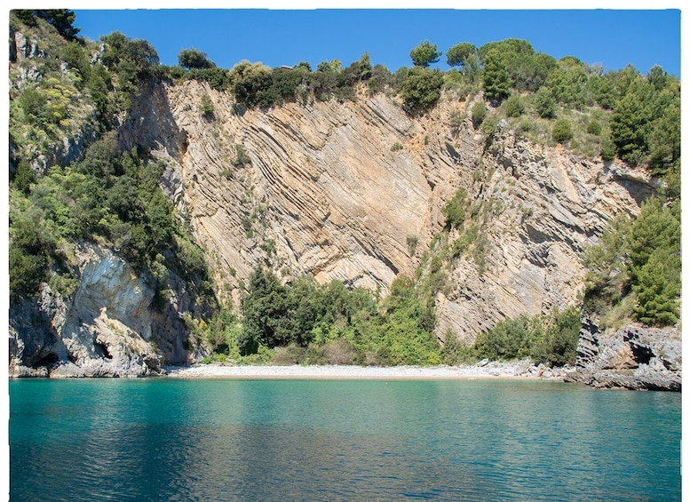 A coastal landscape with a rocky cliff, a clear blue sea, and a sandy beach. 3