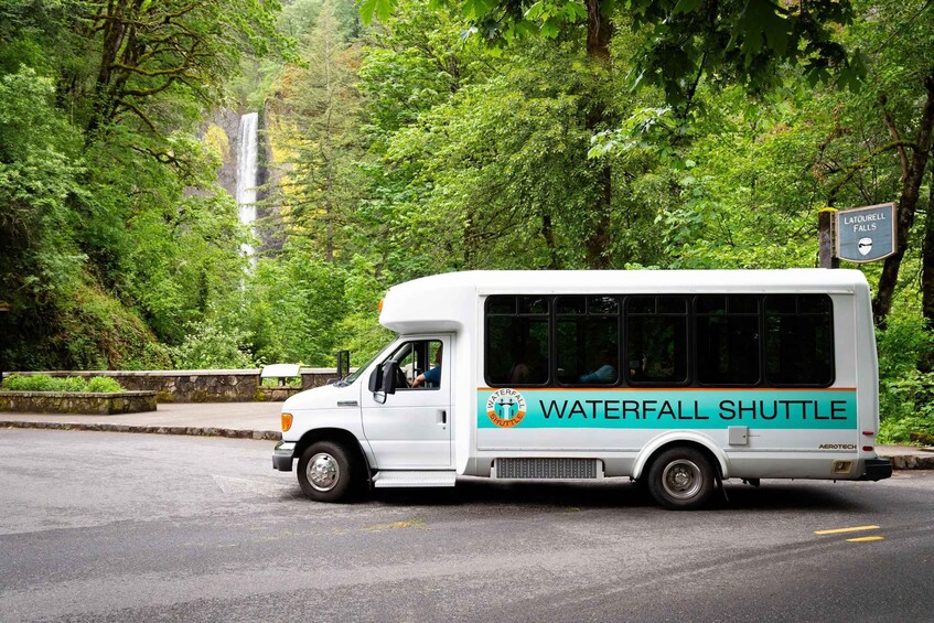 A white shuttle bus with 'Waterfall Shuttle' written on the side, parked on a road with a waterfall in the background. 5