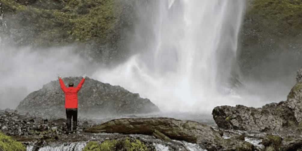 A person standing on a rocky terrain with arms raised, facing a waterfall. 1