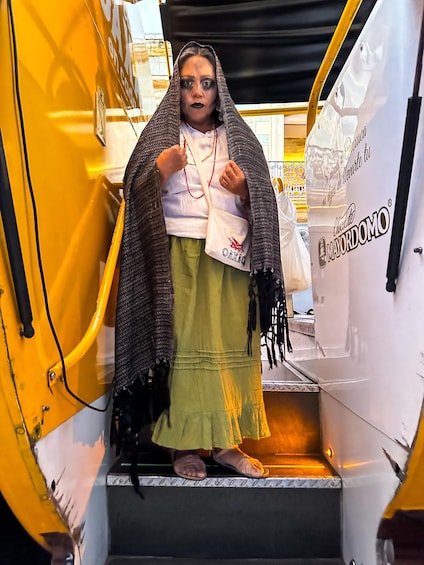 A person standing in the doorway of a yellow vehicle, wearing a shawl, a white shirt, and a green skirt. 2