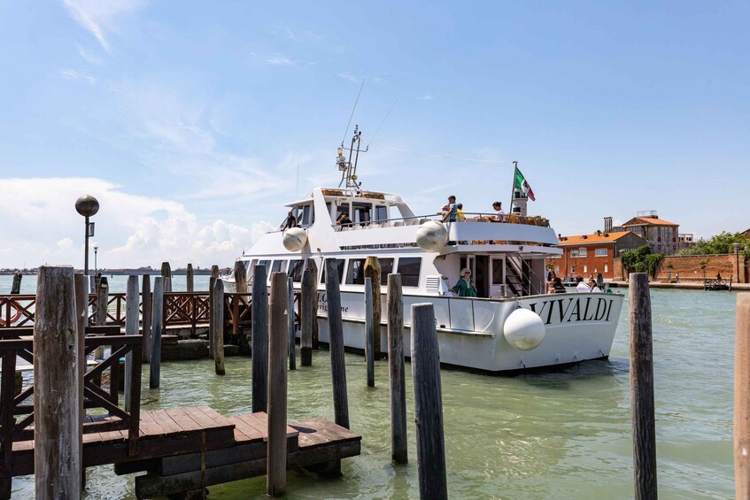 A white boat named 'VIVALDI' docked at a wooden pier with a clear sky and buildings in the background. 1