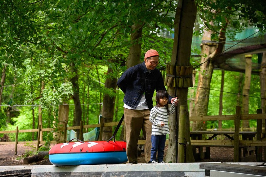 A man and a child near a red and blue inflatable raft in a wooded area with wooden structures. 3