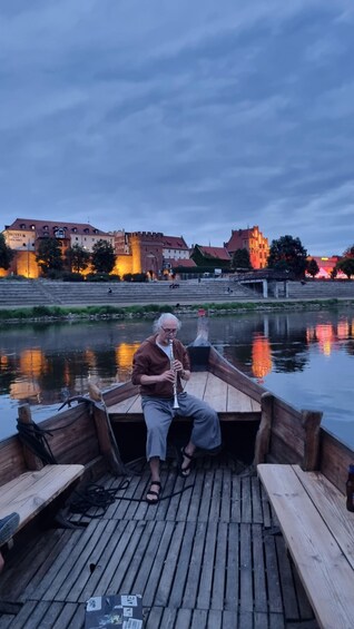 A wooden dock with a person playing a flute. In the background, there are buildings with illuminated windows and a river reflecting lights. 3