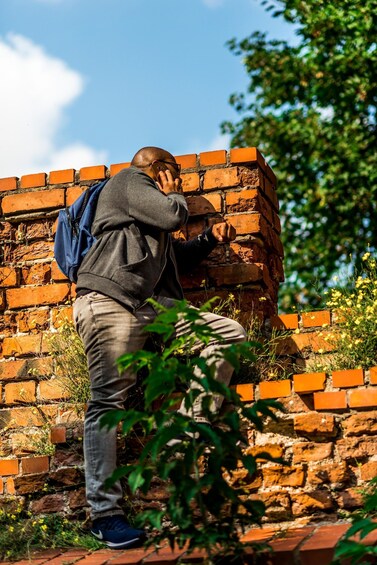 A person taking a photo with a smartphone, standing in front of a brick wall with vegetation around. 2