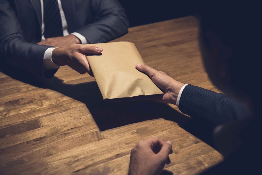 Two individuals exchanging a brown paper bag over a wooden table. 5
