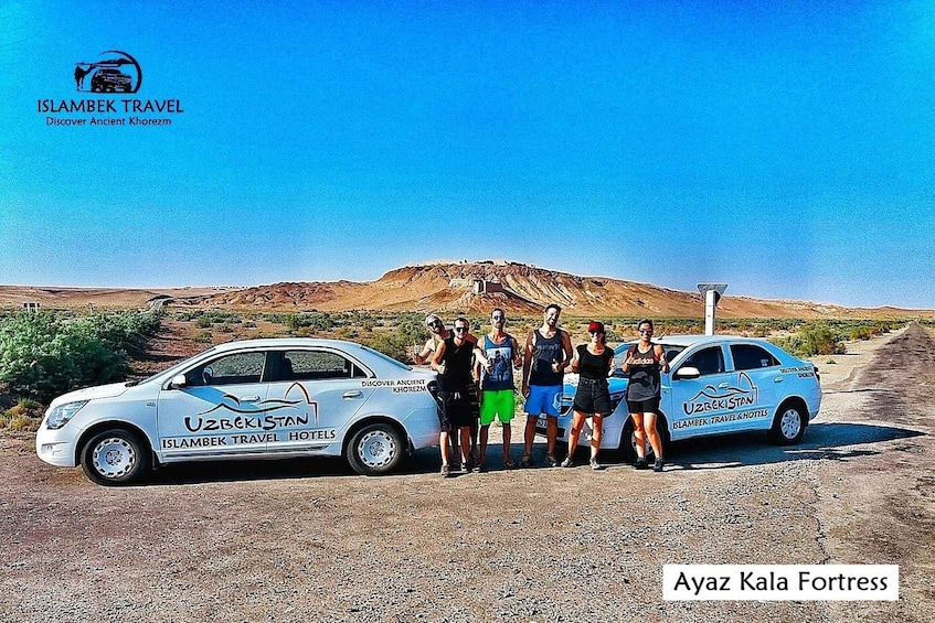 A group of people standing beside two cars with 'Uzbekistan' and 'Islambek Travel Hotels' logos, in front of a barren landscape with a hill in the background. 1