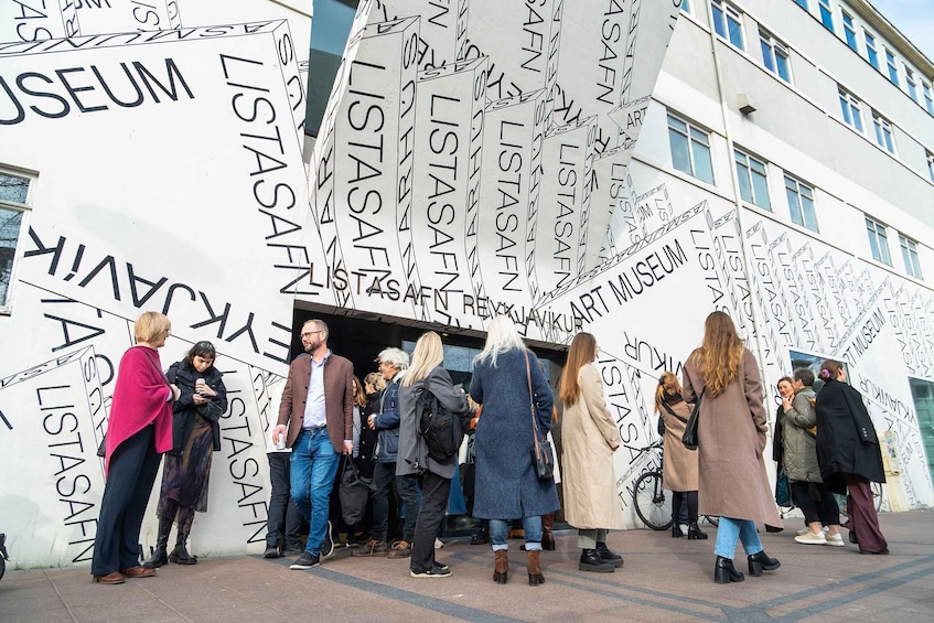 A group of people standing in front of a building with repeated text patterns on the facade. 1