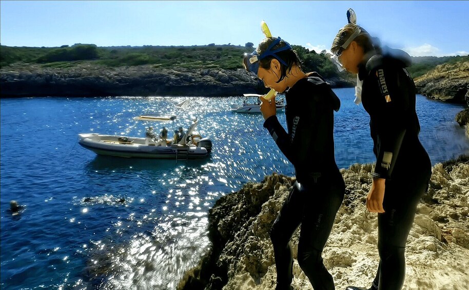 Two people in wetsuits near a rocky shore with a boat in the water. 2