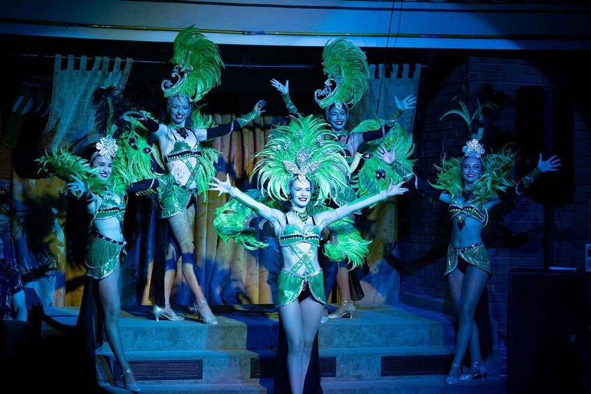 A group of performers in elaborate costumes with green feathers and sequins, dancing on a stage. 1
