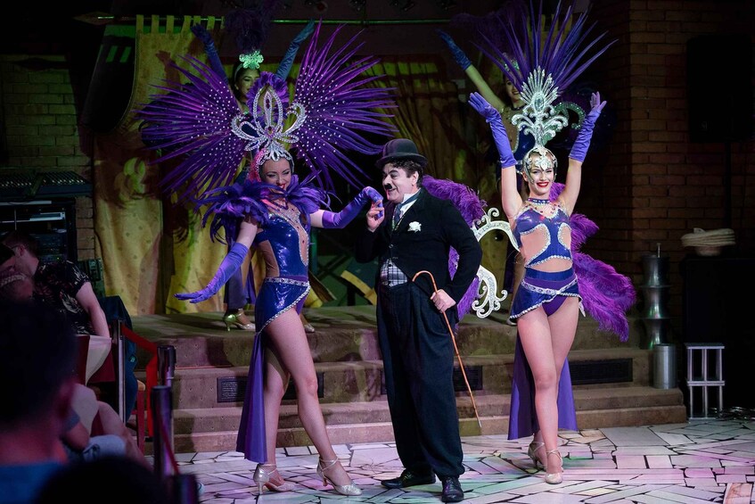 A stage with performers in elaborate costumes, a man in a suit, and a brick wall in the background. 4