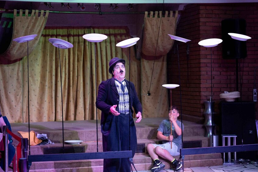 A stage with a performer in a hat and coat, a seated person, and a brick wall in the background. 5