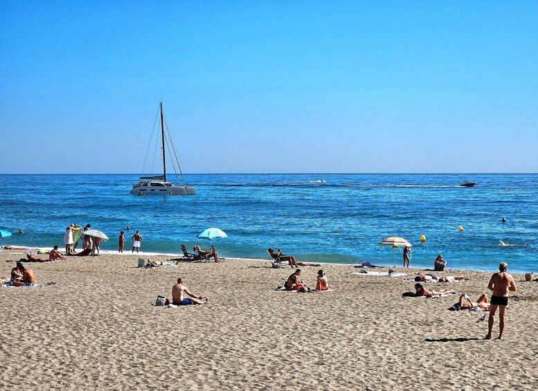A beach with people sunbathing, a sailboat, and a clear blue sky. 2