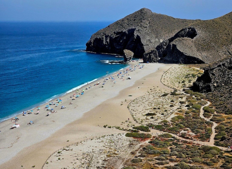 A beach with people sunbathing and walking along the shore, surrounded by rocky cliffs and sparse vegetation. 1