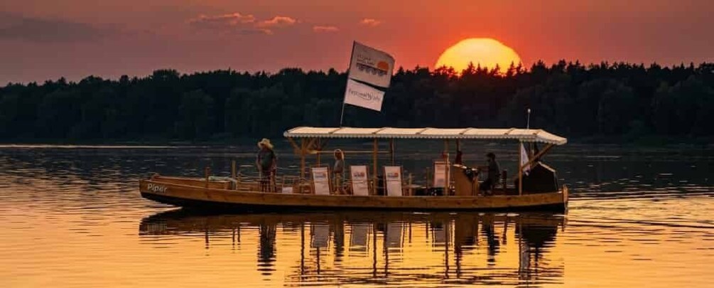 A wooden boat with a covered deck, a flag, and people on a calm lake at sunset. 2