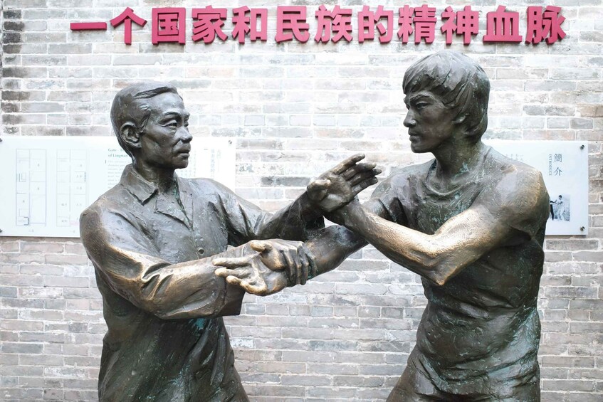 A bronze statue of two figures in a martial arts pose, with a brick wall and informational signs in the background. 5