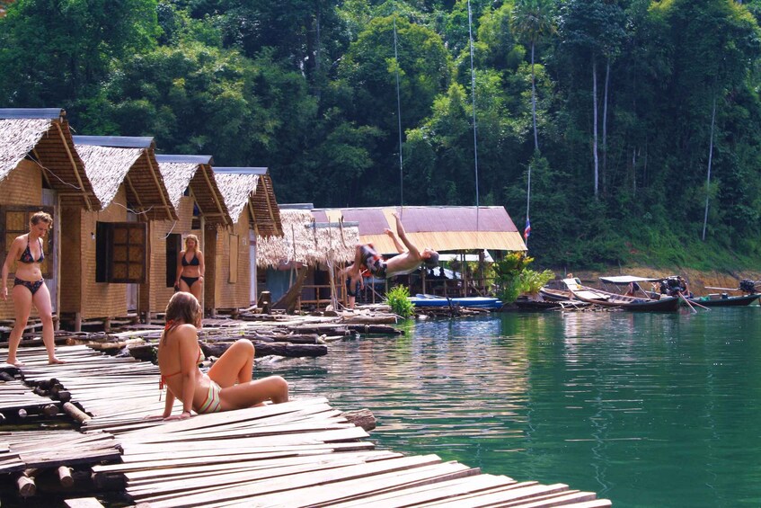 A wooden dock with people relaxing by the water, surrounded by thatched-roof huts and dense forest. 3