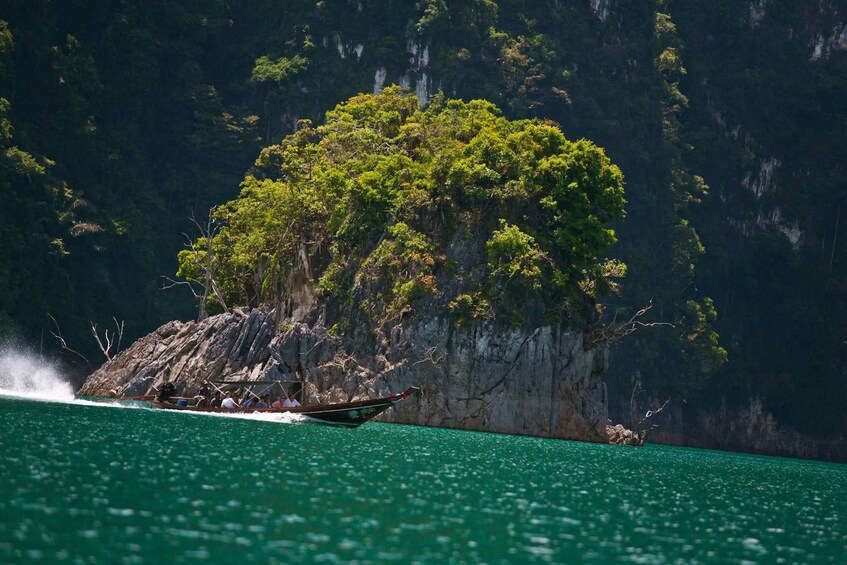 A boat with passengers on a clear day, traveling near a rocky island with dense green foliage. 5