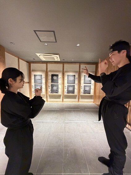 Two people in black martial arts attire practicing in a room with wooden paneling and a tiled floor. 2