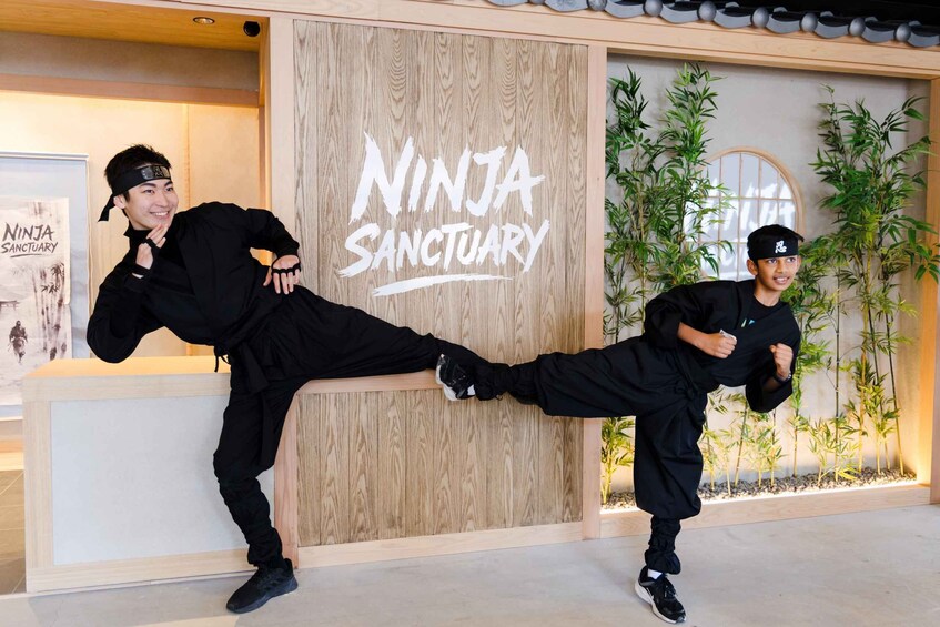 Two individuals performing martial arts kicks in front of a wooden wall with the text 'NINJA SANCTUARY'. 1