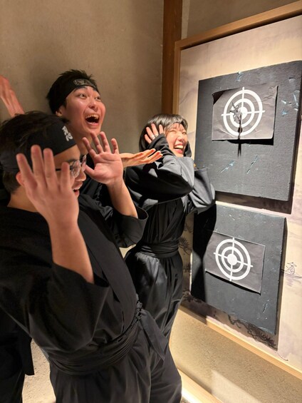 Three individuals in black attire with headbands, standing in front of a wall with target-like drawings. 5