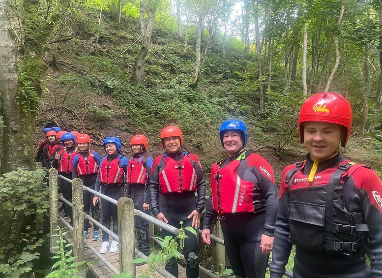 A group of people in red and black gear with helmets, standing on a wooden bridge in a forested area. 1