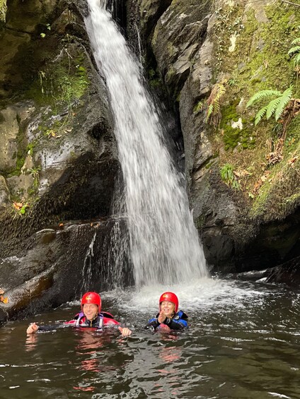 Two people in water near a waterfall surrounded by rocks and greenery. 2