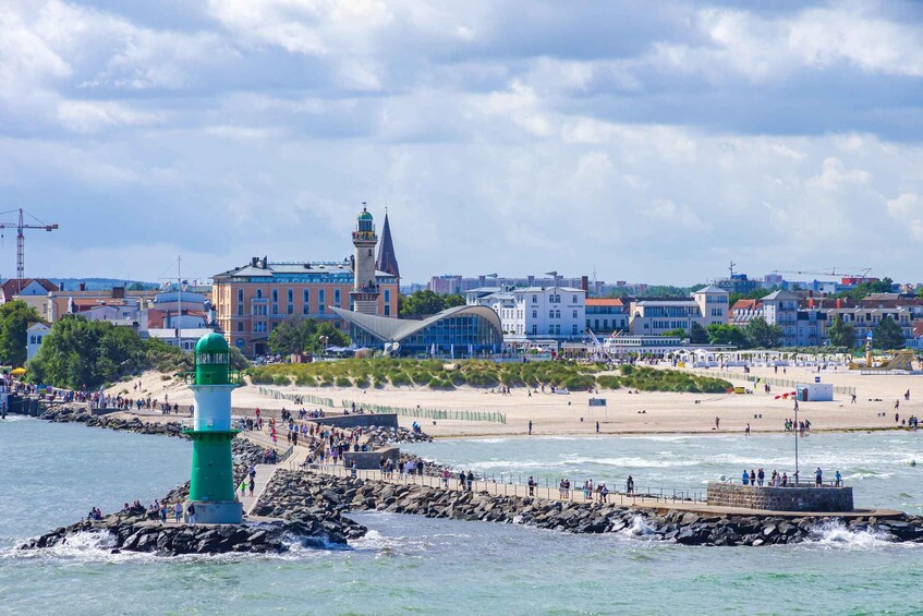 A coastal cityscape with a prominent green lighthouse, a historic building with a tower, and a sandy beach with people. 1