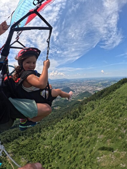 A person in a harness holding onto a paraglider's control bar, with a mountainous landscape and a distant city in the background. 5