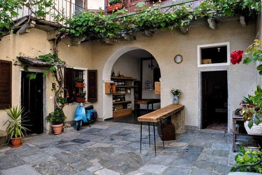 A courtyard with stone flooring, a wooden bench, potted plants, a blue scooter, and a rustic building with an archway. 1