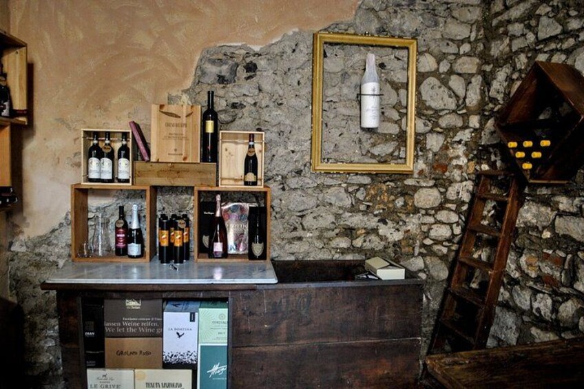 A rustic bar setting with wooden shelves holding bottles, a stone wall, and a framed picture. 3