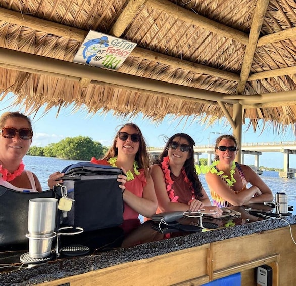 A group of people on a boat with a thatched roof, a sign reading 'GETTIN' FREAKY THE TIKI,' and a view of a bridge and water. 2