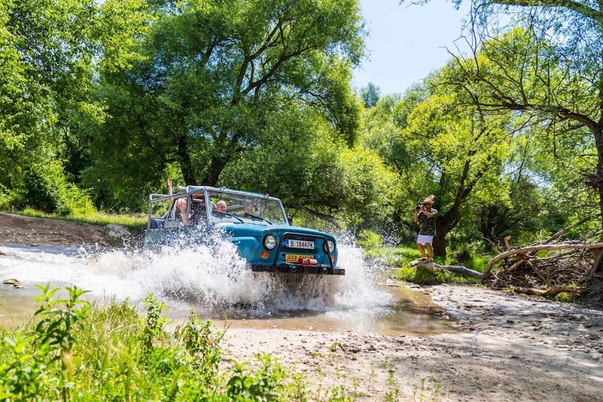 A blue off-road vehicle driving through a shallow water crossing surrounded by lush greenery. 4