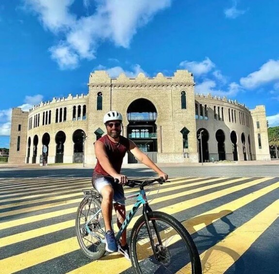 A person on a bicycle in front of a large, historic building with arched windows and a central archway. 5