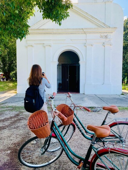 A woman taking a photo with a camera, standing next to a bicycle with baskets. 1