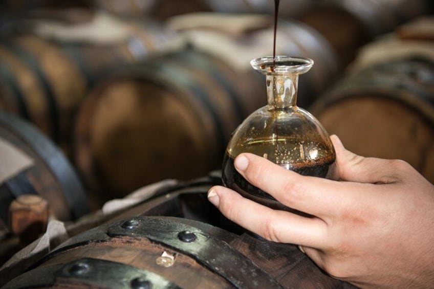 A hand holding a bottle with a dark liquid, barrels in the background. 1