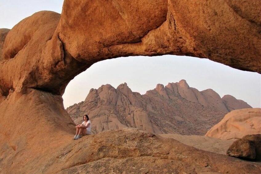 A person sitting under a natural rock arch with a mountainous landscape in the background. 4