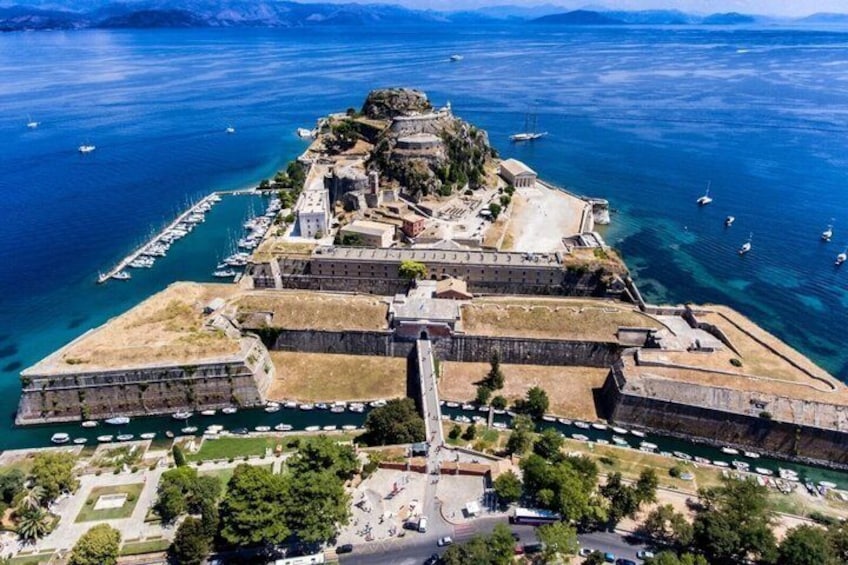 An aerial view of a fortified coastal city with a prominent fortification, a harbor with boats, and a clear blue sea. 1