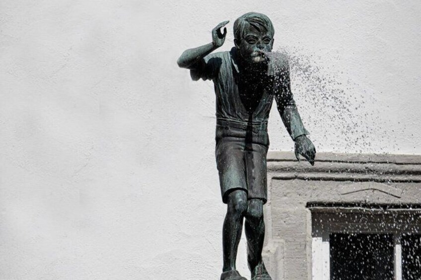 A statue of a person with one hand raised, water splashing around it, set against a plain wall. 4