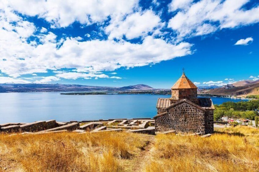 A stone church with a bell tower by a lake, surrounded by dry grass and distant mountains. 1