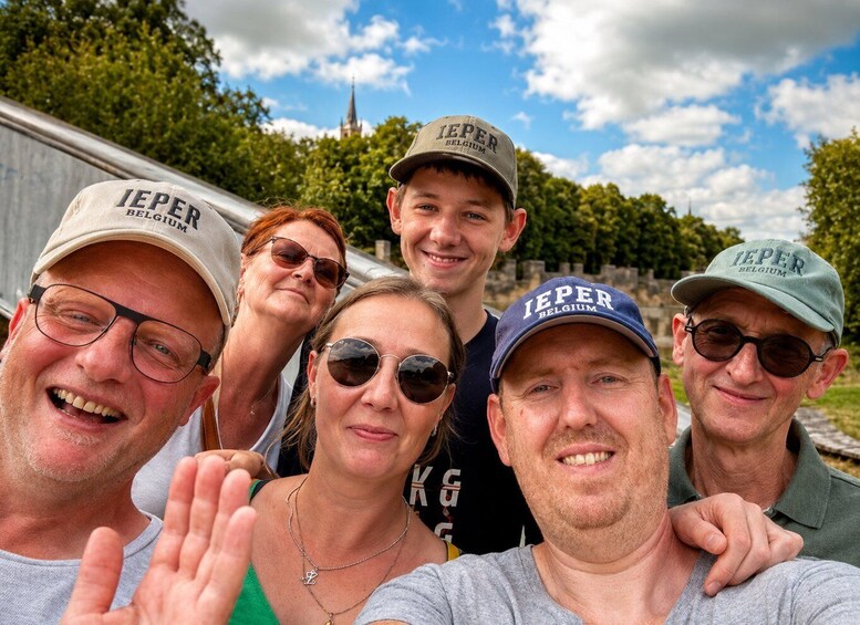 A group of people wearing 'IEPER BELGIUM' caps, standing outdoors with trees and a clear sky in the background. 3