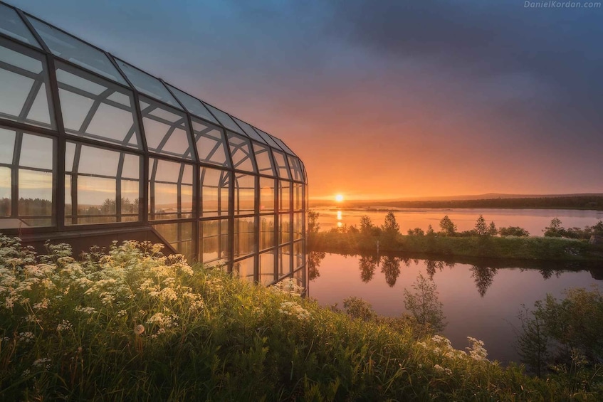 A glass structure reflecting a sunset over a calm lake surrounded by greenery. 1