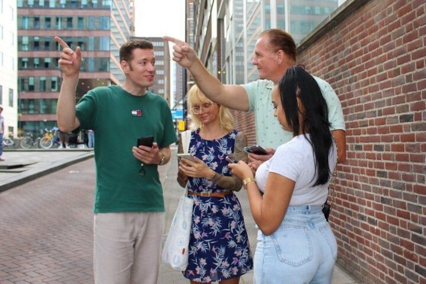 Three people standing on a sidewalk, one pointing and holding a phone, another holding a cup, and a third with a bag. 3