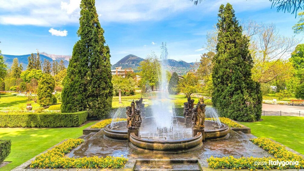 A fountain with statues, surrounded by manicured lawns and tall trees, with mountains in the background. 2