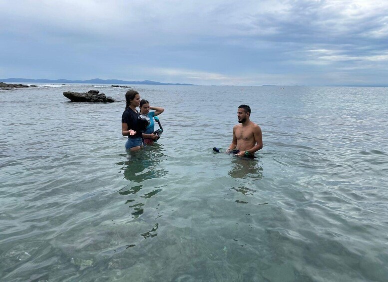 Two people in shallow water, one holding a child, with a rocky shoreline and distant mountains in the background. 2